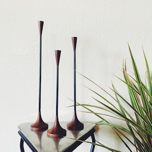 Może przedstawiać: Trzy świeczniki w stylu mid-century modern. Świeczniki mają ciemną drewnianą podstawę i górę z czarnym metalowym trzonkiem. Świeczniki są wystawione na srebrnym stole z czarnymi nogami. W tle roślina z zielonymi i czerwonymi liśćmi.