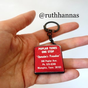 以下が含まれることがあります： 「Poplar Tunes One Stop」と「Operator's Paradise」の文字と住所「308 Poplar Ave. Ph. 525-6348 Memphis, Tenn. 38103」が書かれた白いラベルが付いた赤い金属製のキーホルダー。