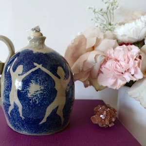 Puede incluir: Una lámpara de aceite de cerámica con un diseño azul y blanco de dos figuras bailando. La lámpara tiene un asa con forma de cinta y está sobre una superficie morada.