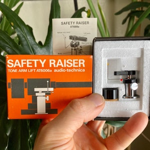 May include: A black and silver tone arm lift for a turntable, labeled "SAFETY RAISER TONE ARM LIFT AT6006a audio-technica".