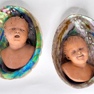Könnte beinhalten: Zwei Terrakotta-Skulpturen von Babyköpfen werden in schillernden Abalone-Muscheln ausgestellt.