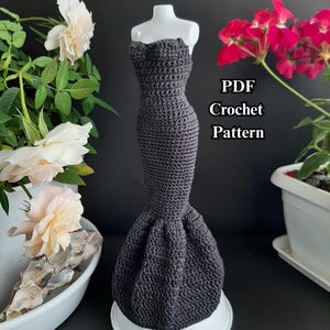 May include: A dark gray crochet dress with a mermaid silhouette, displayed on a white mannequin. The dress has a strapless bodice and a flared skirt. The text "PDF Crochet Pattern" is visible. The background includes white and pink flowers.