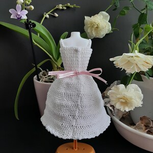 Pode incluir: Um vestido de boneca branco de croch&ecirc; com um la&ccedil;o de fita rosa, exibido em um pequeno suporte laranja. O vestido tem uma bainha com bordas e est&aacute; situado em um fundo de flores e vegeta&ccedil;&atilde;o.