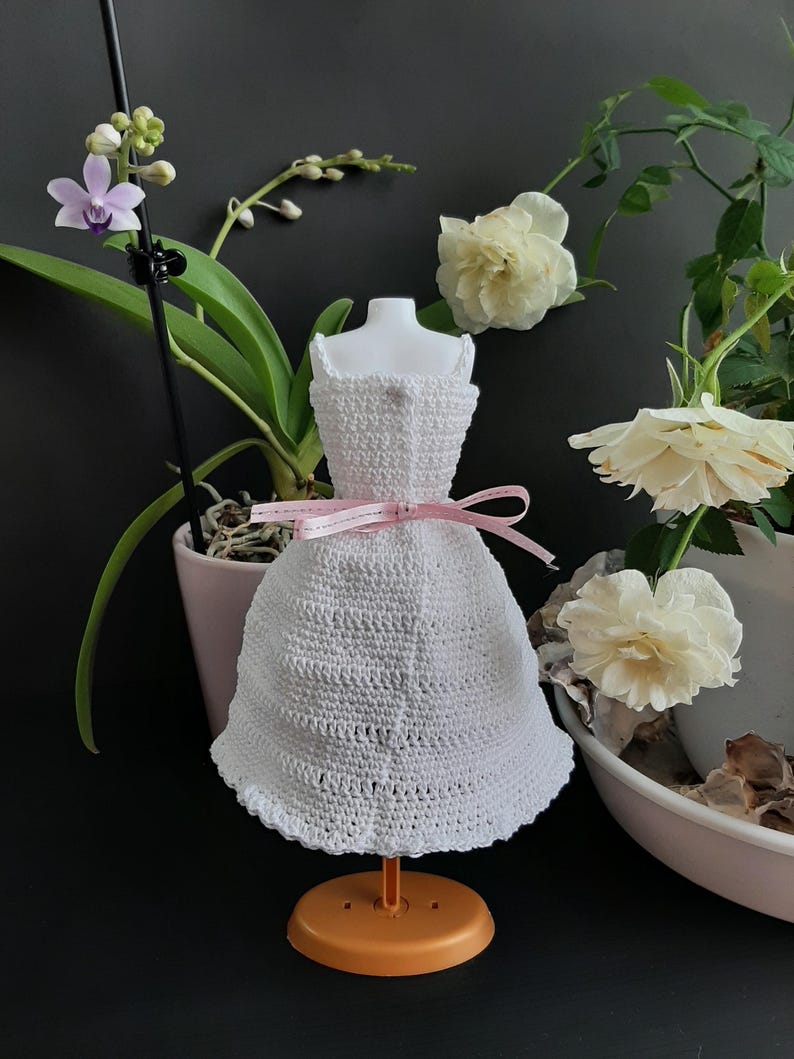 Pode incluir: Um vestido de boneca branco de croch&ecirc; com um la&ccedil;o de fita rosa, exibido em um pequeno suporte laranja. O vestido tem uma bainha com bordas e est&aacute; situado em um fundo de flores e vegeta&ccedil;&atilde;o.