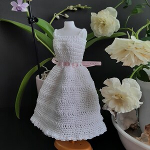Pode incluir: Um vestido branco de croch&ecirc; com um cinto de fita rosa, exibido em um manequim em miniatura. O vestido tem uma bainha com vieiras e &eacute; cercado por rosas brancas e flores de orqu&iacute;deas. O vestido est&aacute; em um pequeno suporte laranja.