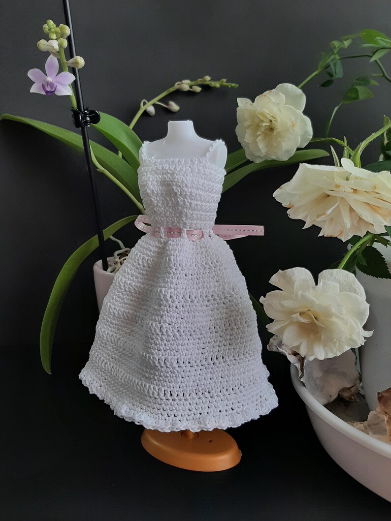 Pode incluir: Um vestido branco de croch&ecirc; com um cinto de fita rosa, exibido em um manequim em miniatura. O vestido tem uma bainha com vieiras e &eacute; cercado por rosas brancas e flores de orqu&iacute;deas. O vestido est&aacute; em um pequeno suporte laranja.