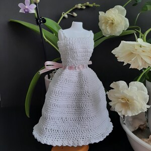 Pode incluir: Um vestido branco de croch&ecirc; em um manequim, com corpete justo e saia rodada. Um cinto de fita rosa marca a cintura. O vestido &eacute; exibido com flores e vegeta&ccedil;&atilde;o, criando uma est&eacute;tica delicada e feminina. Provavelmente &eacute; um vestido em miniatura ou de boneca.
