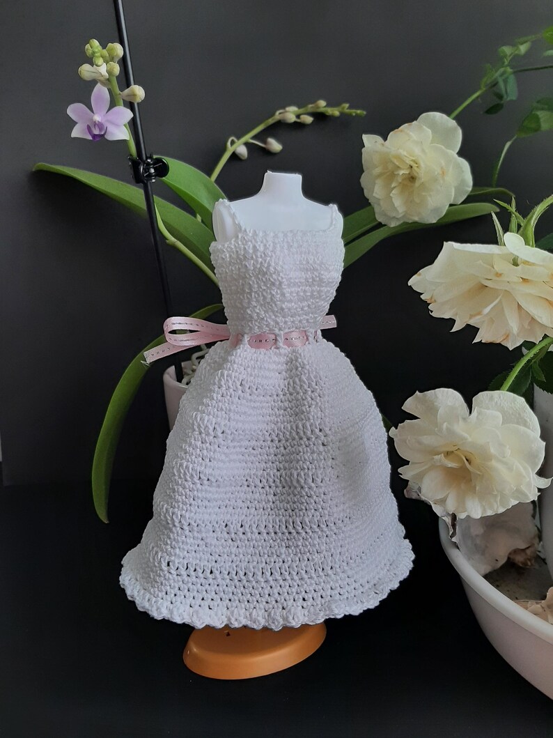 Pode incluir: Um vestido branco de croch&ecirc; em um manequim, com corpete justo e saia rodada. Um cinto de fita rosa marca a cintura. O vestido &eacute; exibido com flores e vegeta&ccedil;&atilde;o, criando uma est&eacute;tica delicada e feminina. Provavelmente &eacute; um vestido em miniatura ou de boneca.