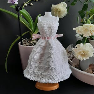 Pode incluir: Um vestido branco de croch&ecirc; em miniatura com um cinto de fita rosa, exibido em um pequeno suporte laranja. O vestido tem um padr&atilde;o detalhado e texturizado e &eacute; projetado para caber em um pequeno manequim. O fundo inclui plantas e flores em vasos.