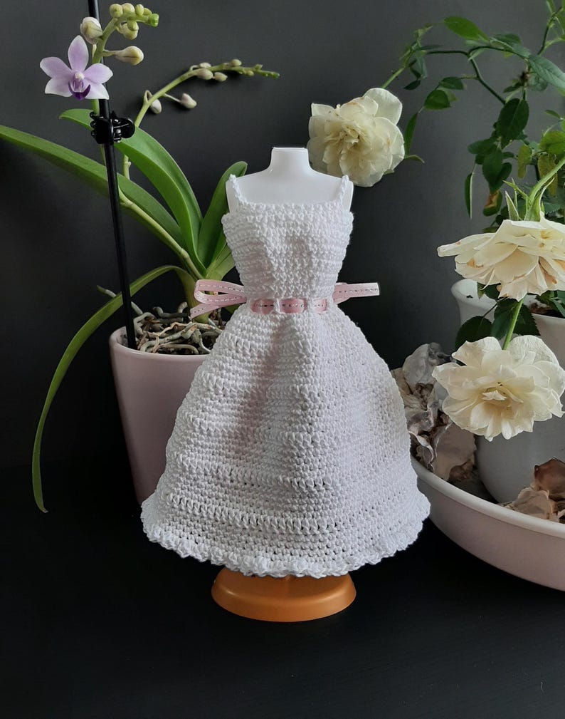 Pode incluir: Um vestido branco de croch&ecirc; em miniatura com um cinto de fita rosa, exibido em um pequeno suporte laranja. O vestido tem um padr&atilde;o detalhado e texturizado e &eacute; projetado para caber em um pequeno manequim. O fundo inclui plantas e flores em vasos.
