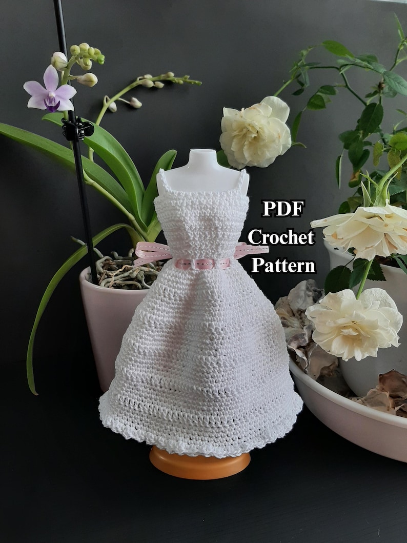 Pode incluir: Um vestido branco de croch&ecirc; exposto em um manequim, com um cinto de fita rosa. O vestido est&aacute; em uma base de madeira. O texto &agrave; direita diz "PDF Crochet Pattern". O fundo inclui flores e plantas.