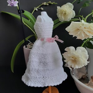 Pode incluir: Um pequeno vestido branco de croch&ecirc; exibido em um suporte laranja. O vestido tem um padr&atilde;o texturizado e &eacute; adornado com uma fita rosa. O fundo inclui flores brancas e uma orqu&iacute;dea em vaso.