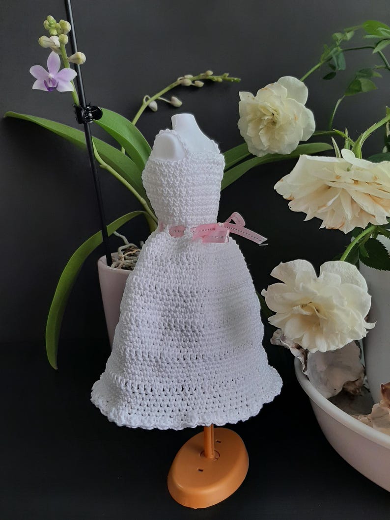Pode incluir: Um pequeno vestido branco de croch&ecirc; exibido em um suporte laranja. O vestido tem um padr&atilde;o texturizado e &eacute; adornado com uma fita rosa. O fundo inclui flores brancas e uma orqu&iacute;dea em vaso.
