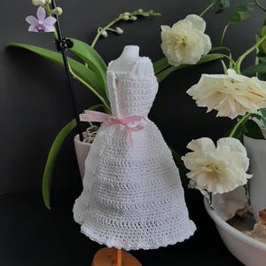 Pode incluir: Um vestido branco de croch&ecirc; em um manequim, com uma fita rosa amarrada na cintura. O vestido &eacute; exibido em um suporte laranja. Rosas brancas e uma orqu&iacute;dea no fundo.