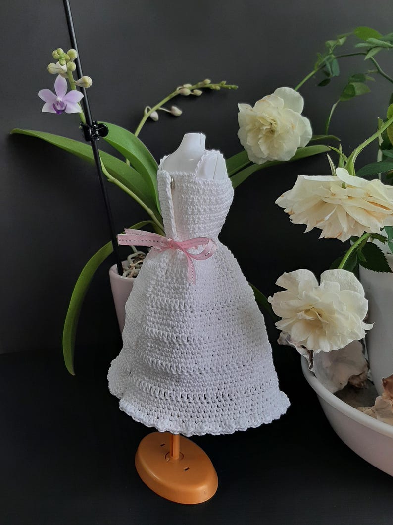 Pode incluir: Um vestido branco de croch&ecirc; em um manequim, com uma fita rosa amarrada na cintura. O vestido &eacute; exibido em um suporte laranja. Rosas brancas e uma orqu&iacute;dea no fundo.