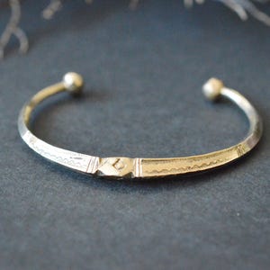 Op de afbeelding: Een zilverkleurige metalen armband met een centrale ruitvormige gravure, geflankeerd door geëtste golvende lijnpatronen. De armband heeft een slank, open ontwerp met kleine, afgeronde bolvormige uiteinden aan beide kanten.