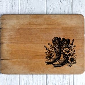 May include: A wooden cutting board with a decorative design. The design features a pair of cowboy boots with floral and wheat accents. The boots are black and the flowers are sunflowers. The cutting board is light brown.