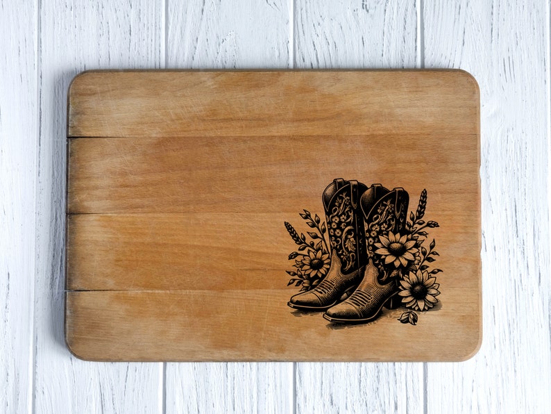 May include: A wooden cutting board with a decorative design. The design features a pair of cowboy boots with floral and wheat accents. The boots are black and the flowers are sunflowers. The cutting board is light brown.