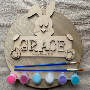Può includere: Un cartello di legno a forma di coniglio con il nome "GRACE" e il testo "Buona Pasqua 2024". Il cartello è accompagnato da sei piccoli barattoli di vernice e due pennelli.