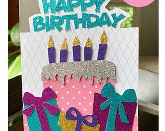 May include: A birthday card with a layered cake design. The cake is decorated with purple candles and silver glitter. The card also features presents with bows and the words "Happy Birthday" in turquoise letters.
