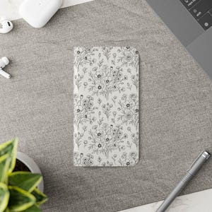 May include: A floral phone case featuring a delicate black and white botanical pattern. The design includes detailed line drawings of flowers and leaves, creating a classic and elegant look. The case is rectangular and appears to be made of a smooth material.