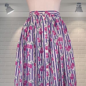 Op de afbeelding: Een knooprok met een verticaal streepjespatroon in wit en paars. De rok is versierd met een herhalend patroon van roze kersen met groene bladeren. De rok wordt op een mannequin getoond.