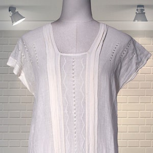 May include: A white, short-sleeved blouse with a square neckline. The blouse features vertical pleats and decorative stitching down the front. The bottom hem is gathered with a tie detail. The garment is displayed on a mannequin.