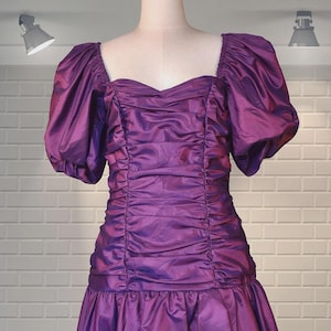 May include: A vintage purple dress with a sweetheart neckline and short, puffed sleeves. The bodice is ruched, and the skirt flares out from the waist. The dress is made of a shiny fabric and is displayed on a mannequin.