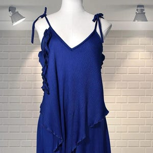 以下が含まれることがあります： Vネックと結びストラップが付いた鮮やかなブルーのドレス。 ボディスはプリーツデザインで、フリルオーバーレイが付いています。 ドレスは流れるようなスカートで、マネキンに展示されています。 背景は白いレンガの壁です。