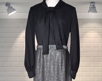 May include: Black long-sleeved dress with a silver, glittery skirt. The dress has a tie-neck detail and a matching belt with a rectangular buckle. The dress is displayed on a mannequin against a white brick wall.