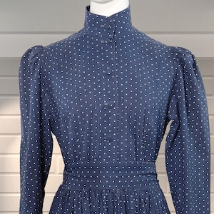 May include: A navy blue dress with white polka dots, long sleeves, and a high collar. The dress features a fitted waist, a button-down front, and a flared skirt. The dress is displayed on a mannequin against a white brick wall.