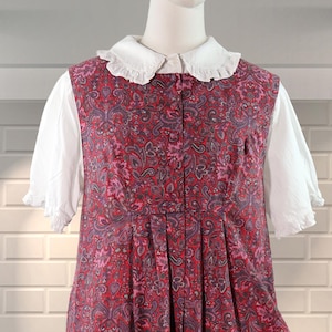 以下が含まれることがあります： ピンクとグレーの色合いのペイズリー柄の赤いロングドレス。 ドレスは白いフリルカラーと半袖です。 ドレスは白いレンガの壁を背景にマネキンに展示されています。