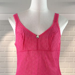 May include: A vibrant pink slip dress displayed on a mannequin. The dress has a V-neck, lace detailing, and a small decorative element at the bust. The fabric has a textured pattern throughout.