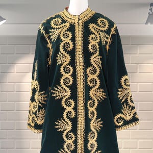 Peut inclure: Long manteau en velours vert foncé avec broderies dorées complexes. Les broderies présentent des motifs floraux et enroulés ornés sur le devant, les manches et le col. Le manteau est présenté sur un mannequin devant un mur de briques blanches.