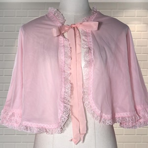 May include: A pale pink cape with lace edging and a bow at the neck, displayed on a mannequin. The cape's fabric is sheer and lightweight, with a vintage style. The lace trim adds a delicate touch to the garment's design.