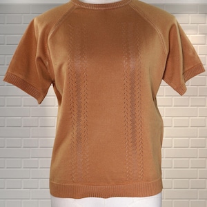 May include: A tan-coloured knitted top with short sleeves and a crew neck. It has ribbed cuffs and vertical openwork detailing down the front. The garment is displayed on a mannequin against a white brick wall.