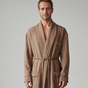 May include: A long, tan-colored robe featuring a shawl collar, long sleeves, and two front pockets. The garment is secured at the waist with a matching braided cord tie that includes tassel ends. Darker brown decorative stitching accents the collar, cuffs, and pockets.