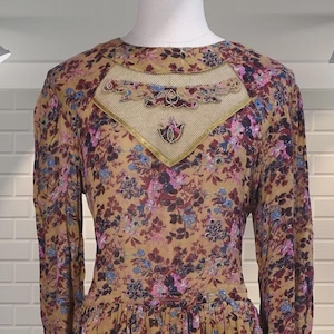 Könnte beinhalten: Langärmliges Kleid mit floralem Muster in Rosa-, Blau- und Lilatönen auf beigem Hintergrund. Das Kleid hat einen dekorativen Ausschnitt und eine geraffte Taille, die eine fließende Silhouette erzeugt.