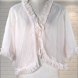 May include: A sheer, white, short-sleeved bolero jacket with ruffled trim along the edges and neckline. The jacket is displayed on a mannequin against a white brick wall background.