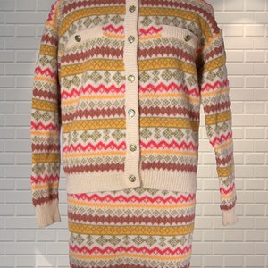 May include: A vintage two-piece knit set featuring a cardigan and a skirt. The set is cream-colored with a geometric pattern in shades of red, yellow, green, and brown. The cardigan has gold buttons and two small pockets.