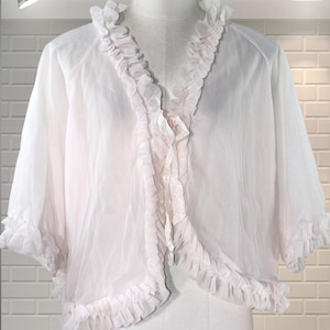 May include: A sheer, white, ruffled bolero jacket displayed on a mannequin. The jacket features a V-neckline and short sleeves, both trimmed with delicate ruffles. The fabric appears lightweight and flowing, creating a soft, feminine silhouette.