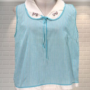 May include: A sleeveless top with vertical turquoise and white stripes. It has a white Peter Pan collar with embroidered pink flowers and a turquoise tie. The hem is white with a turquoise trim.