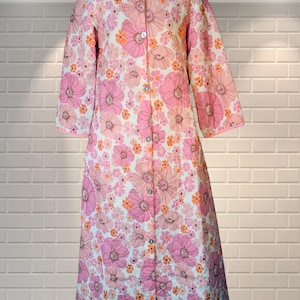 Peut inclure: Une robe de chambre à manches longues avec une base blanche et un motif floral dense dans des tons de rose, magenta et orange. Le vêtement présente un col arrondi, une fermeture boutonnée sur toute la longueur et un liseré rose.