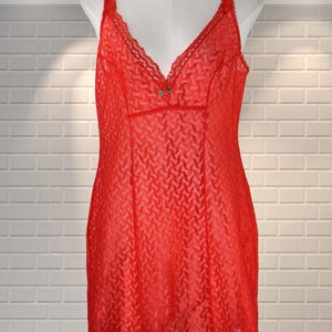 May include: A red slip dress with a V-neck and lace detailing. The dress has a textured pattern and is displayed on a mannequin. The garment is sleeveless and has a small decorative element at the bust.