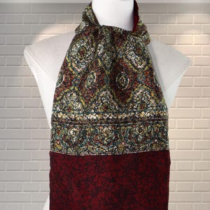 May include: A patterned scarf with a mix of colors, including red, green, and yellow, is draped over a mannequin. The scarf has a detailed design and a fringed bottom edge. The top portion features a complex pattern, while the bottom is a solid deep red.