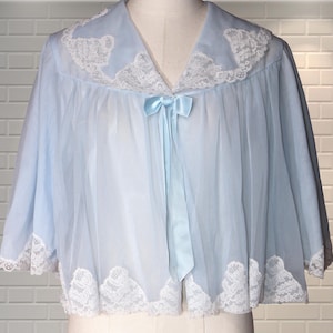 Puede incluir: Una capa corta de color azul claro semitransparente con un cuello ancho estilo marinero adornado con encaje floral blanco. La prenda tiene mangas cortas y holgadas, un cuerpo fruncido y un lazo de satén azul claro a juego en el cuello.
