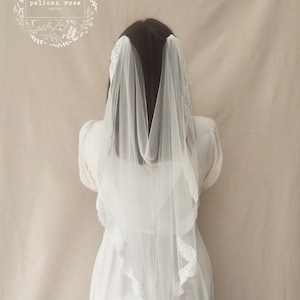 May include: A white bridal veil with intricate lace edging, worn by a person in a white dress. The veil is transparent, revealing the dress beneath. The image has a soft, romantic feel, with the text "pelican rose" inside a floral wreath.