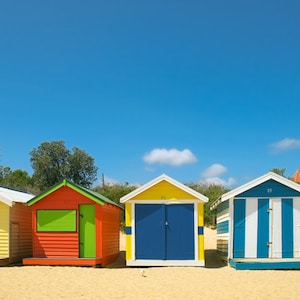Könnte beinhalten: Eine Reihe bunter Strandhütten an einem Sandstrand unter einem strahlend blauen Himmel. Die Hütten sind in verschiedenen Farben wie Gelb, Rot, Blau und Grün mit weißen Akzenten bemalt. Das Bild wurde an einem sonnigen Tag aufgenommen.