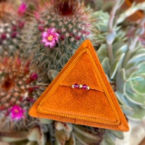 Może przedstawiać: Złoty pierścionek z trzema małymi, czerwonymi, kwadratowymi kamieniami szlachetnymi ułożonymi w rzędzie. Pierścionek jest prezentowany na trójkątnym, pomarańczowym aksamitnym poduszce.