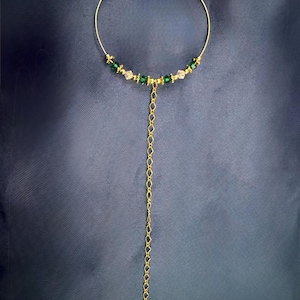 May include: A gold-coloured necklace with a delicate design. The necklace features a circular upper portion adorned with green and clear beads. A long chain of gold-coloured links dangles from the centre, creating a Y-shaped pendant. The background is a dark blue fabric.